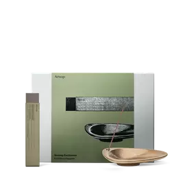 Una varilla de incienso reposa en un soporte color bronce junto a un estuche alto, ante una caja de regalo Aesop verde oliva.