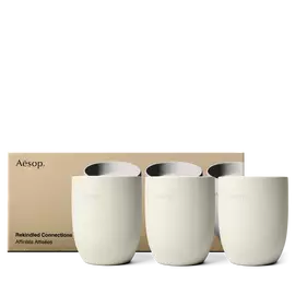 Tres recipientes de velas Aesop color crema se alinean frente a una caja dorada impresa con imagenes de las velas correspondientes.