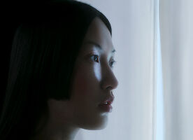 Un perfil cinematografico en primer plano de una mujer con cabello oscuro y liso y piel clara, mirando pensativa hacia una fuente de luz suave y fria.