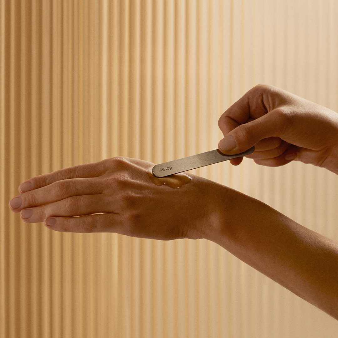 Una persona usa una espatula de Aesop para aplicar un producto de cuidado facial en el dorso de la mano.