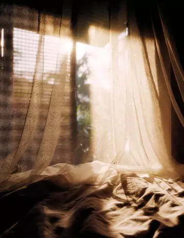 Cortinas vaporosas, dispuestas sobre una tela arrugada junto a una ventana iluminada por el sol, revelan tonos beige calidos, sombras suaves y pliegues translucidos.