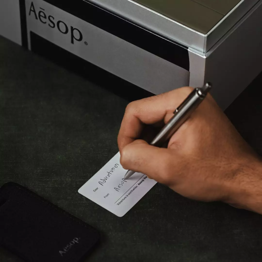 Una mano escribe en una pequena tarjeta blanca junto a una etiqueta de equipaje negra de Aesop y un estuche plateado de Aesop sobre tela verde oscuro.