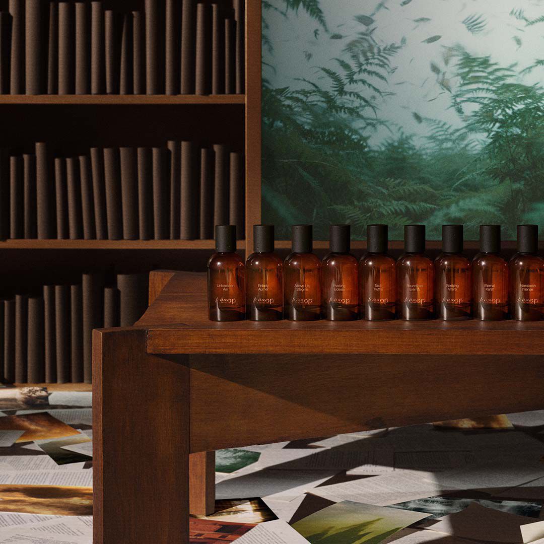 Una fila de frascos de perfume de vidrio ambar de Aesop exhibidos en un estante de madera frente a una biblioteca y una obra de arte verde.