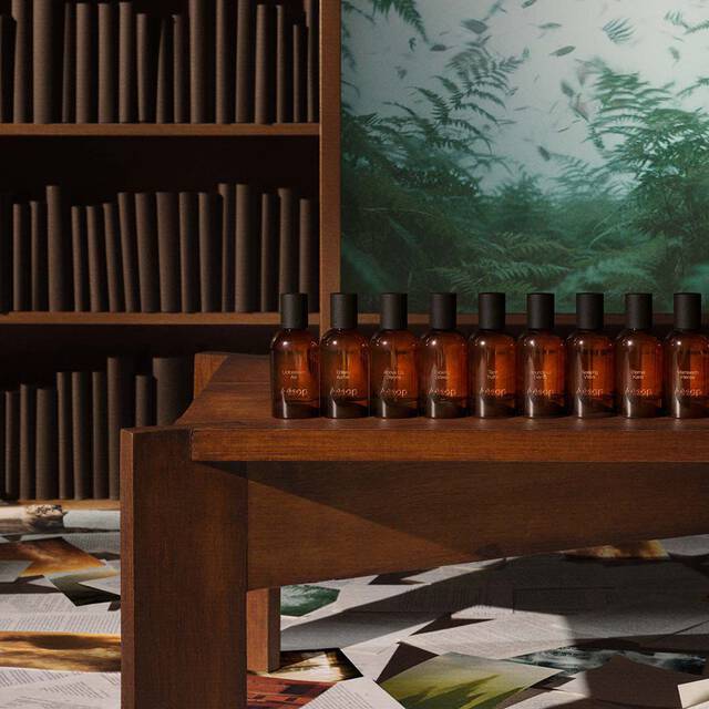 Una fila de frascos de perfume de vidrio ambar de Aesop exhibidos en un estante de madera frente a una biblioteca y una obra de arte verde.