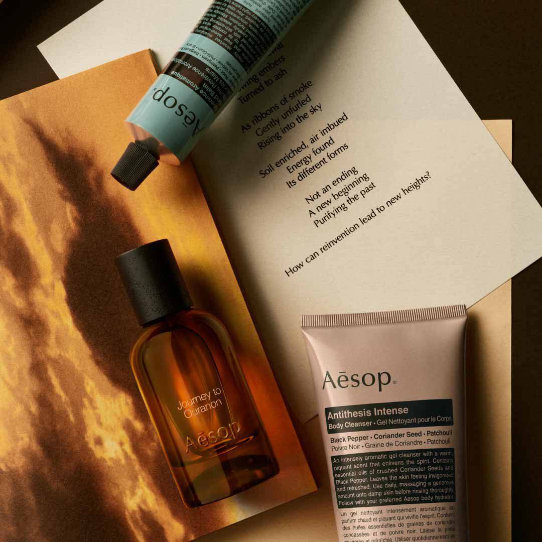Frasco de Aesop Journey to Ouranon con dispensador y tubo, dispuestos sobre papeles con una imagen de paisaje luminoso.