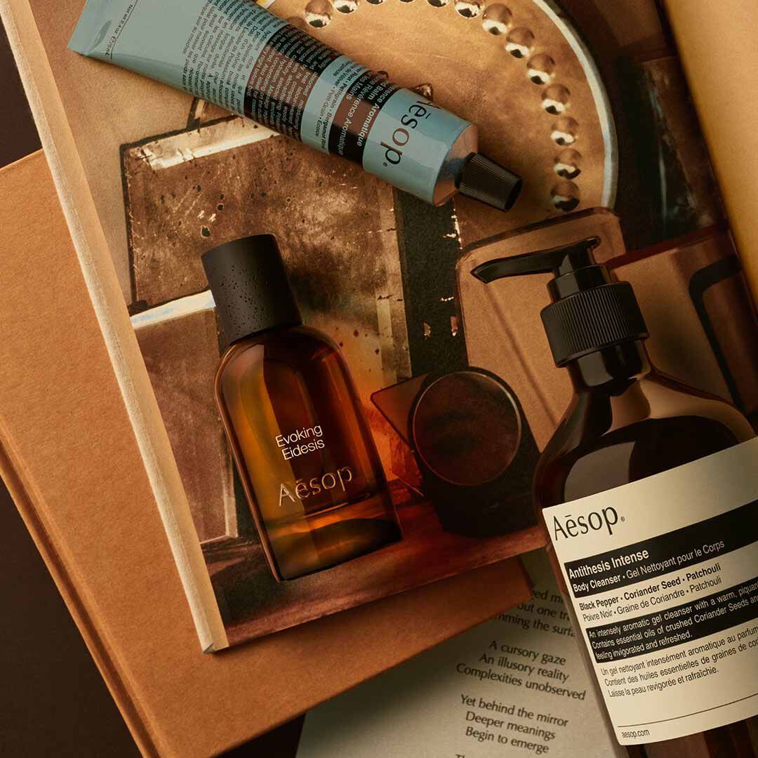 Frasco de Aesop Evoking Eidesis con dispensador y tubo azul, dispuestos sobre papeles e imagenes de una bandeja espejada.