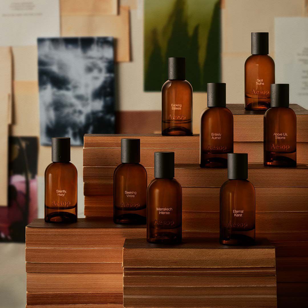 Coleccion de frascos de fragancia ambarina dispuestos sobre bloques de madera, ante laminas y papeles superpuestos en suaves tonos crema y carbon.