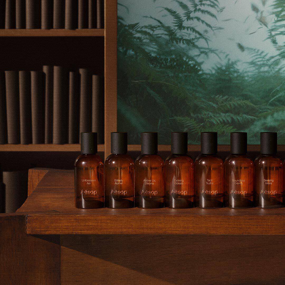 Fila de frascos ambar de fragancias Aesop sobre una mesa de madera, junto a estantes y una imagen de un helecho verde.