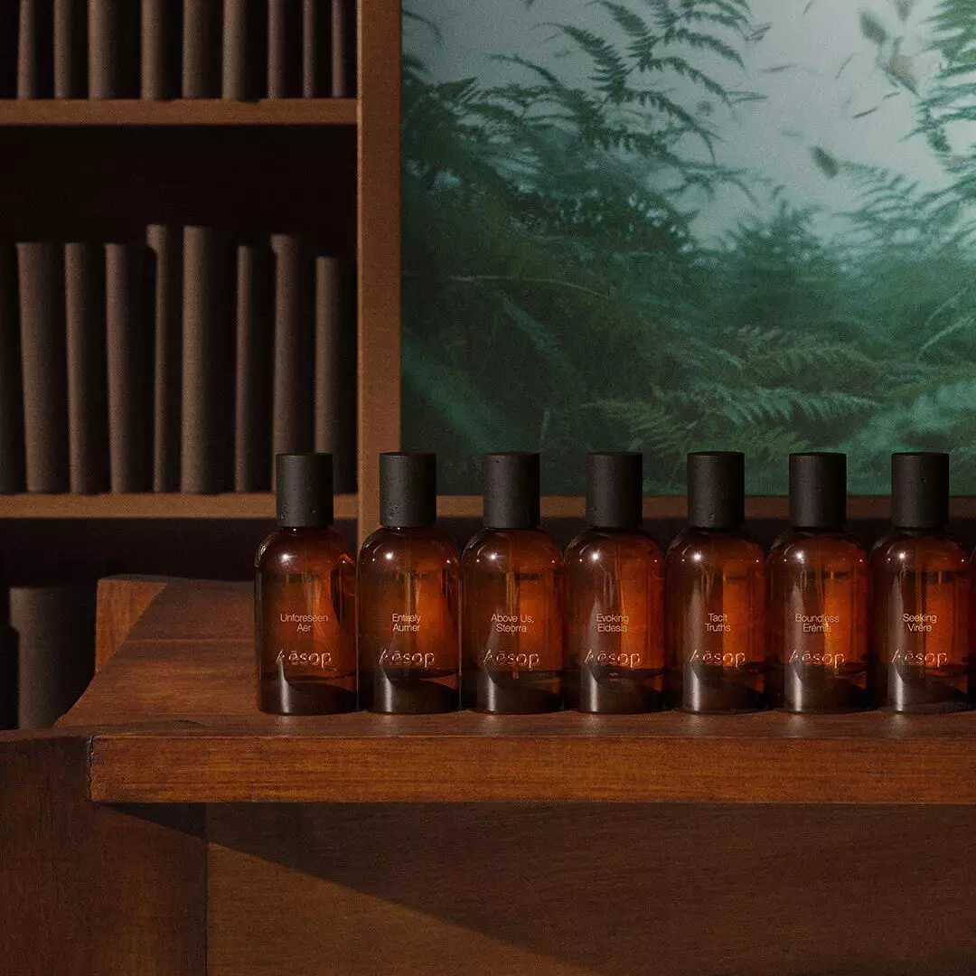 Fila de frascos ambar de fragancias Aesop sobre una mesa de madera, junto a estantes y una imagen de un helecho verde.
