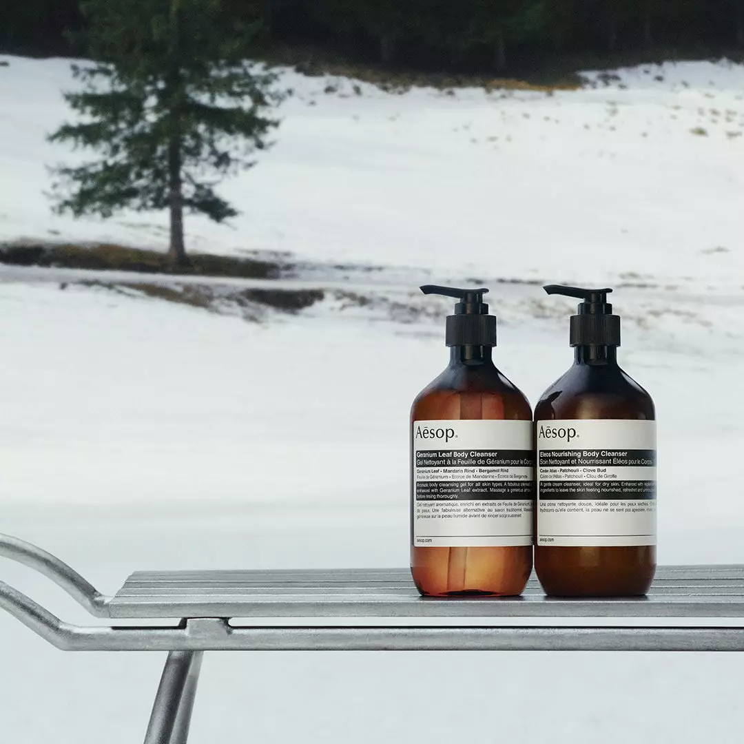Two amber Aesop bottles with black pumps and white labels stand side by side on a metal sledge in the snow.