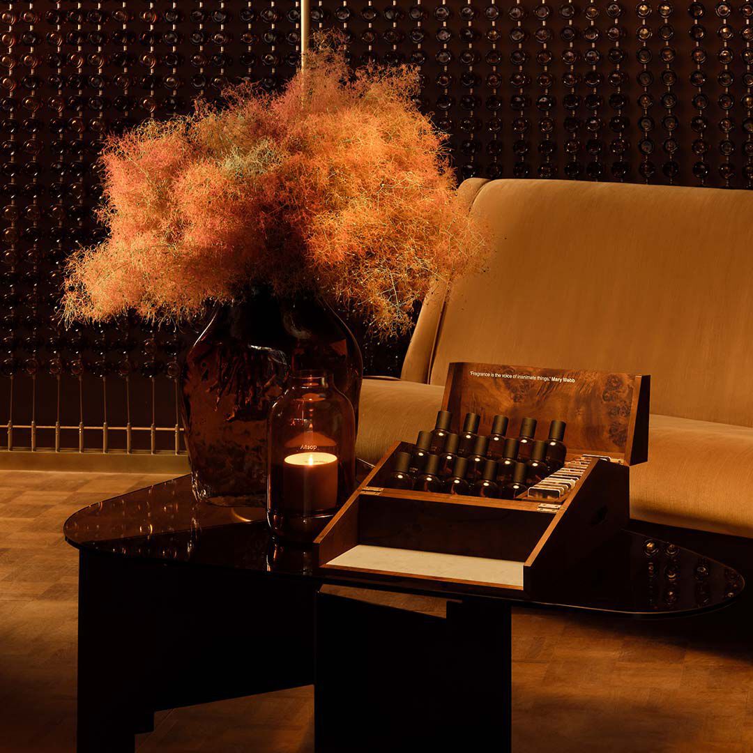 Bodegon interior en tonos ambar con un sofa, una mesa oscura, un arreglo floral seco y frascos de fragancia expuestos en una caja de presentacion abierta.
