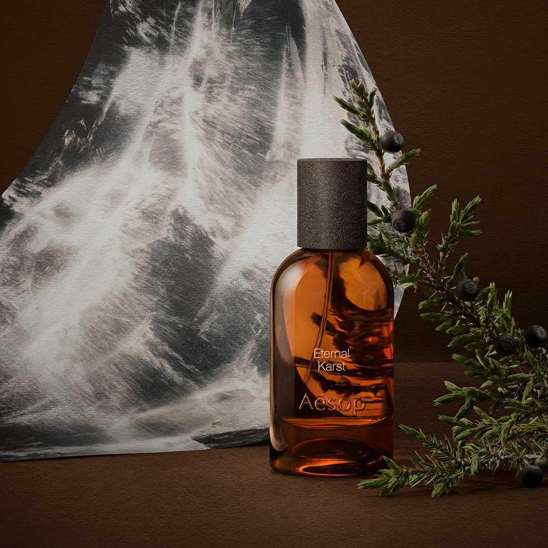 Frasco ambar de fragancia Aesop junto a ramitas de romero y una imagen monocromatica de agua, sobre una superficie marron oscuro.