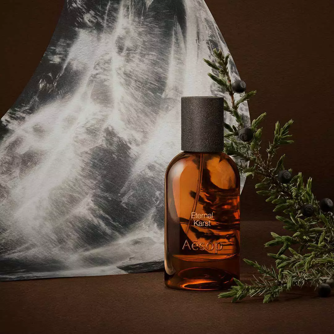 Frasco ambar de fragancia Aesop junto a ramitas de romero y una imagen monocromatica de agua, sobre una superficie marron oscuro.