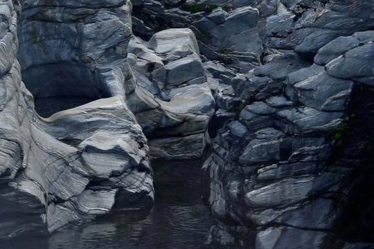 Layered slate-grey rock formations above dark water, showing striated mineral textures, cracks, and naturally sculpted contours.
