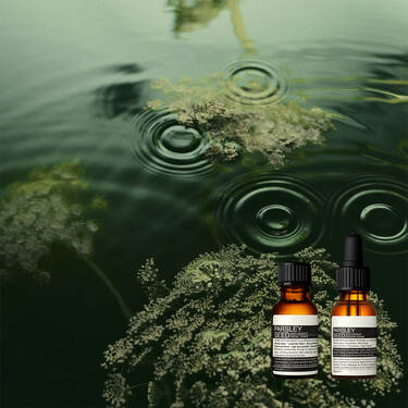 Dos frascos goteros de color ambar de Aesop sobre musgo verde sumergido bajo el agua oscura y ondulante.
