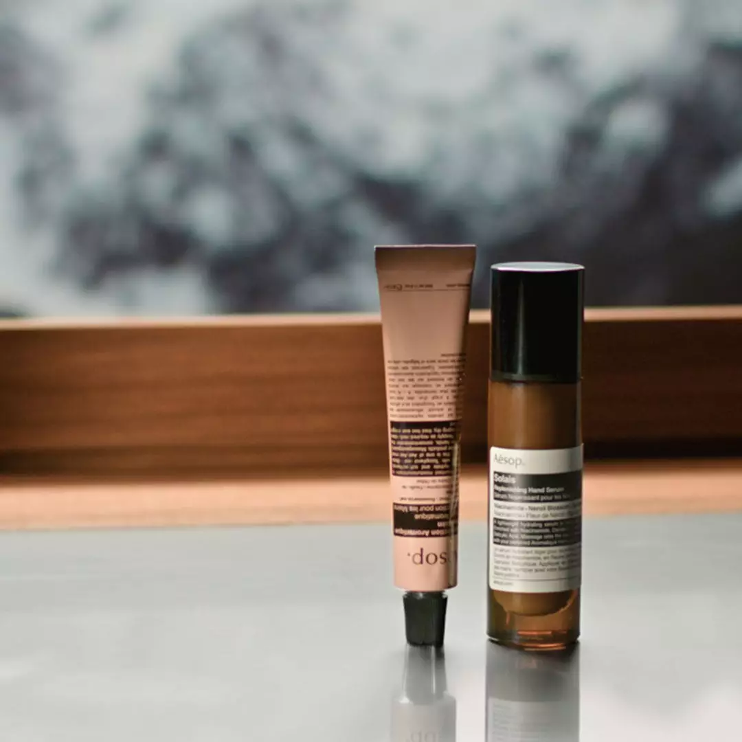 A pink Aesop tube and a slim amber bottle with a black cap stand on a reflective surface beside a wooden-framed window.