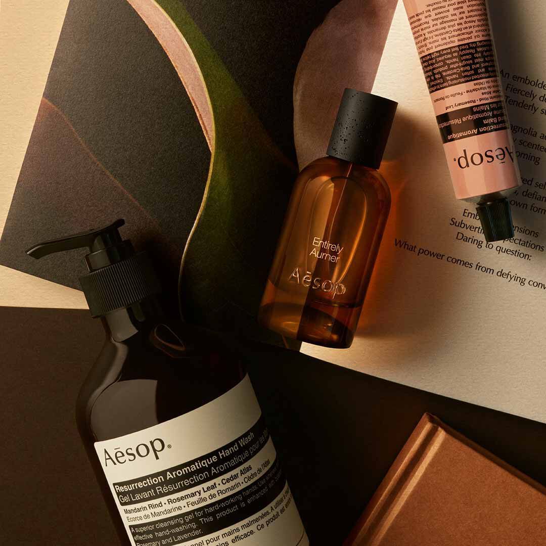 Frasco de Aesop Entirely Aurner con dispensador y tubo color melocoton, dispuestos sobre papeles y formas oscuras curvadas.