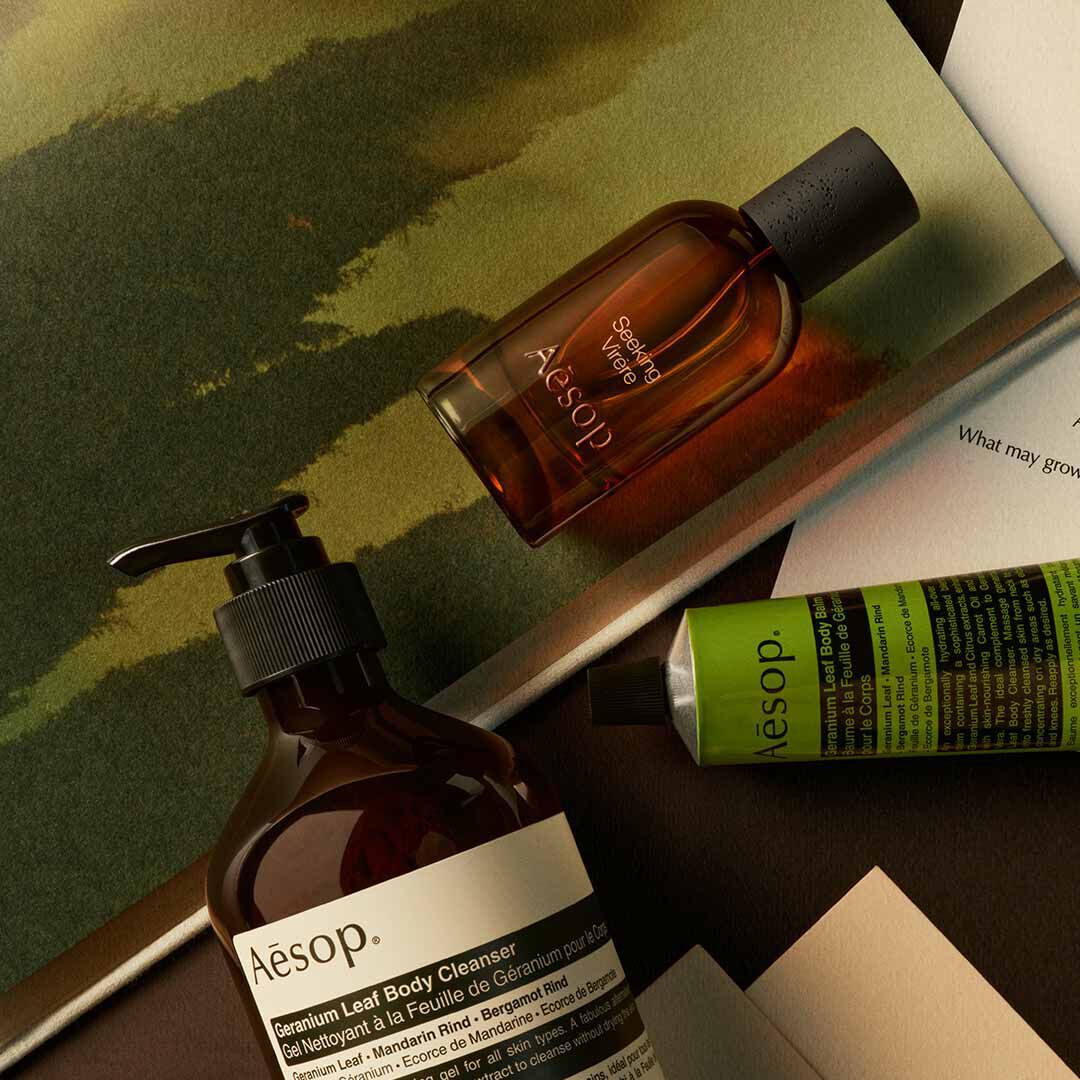 Frasco de Aesop Seeking Virere con dispensador y tubo verde, dispuestos sobre papeles con imagenes pintadas en tonos verdes.