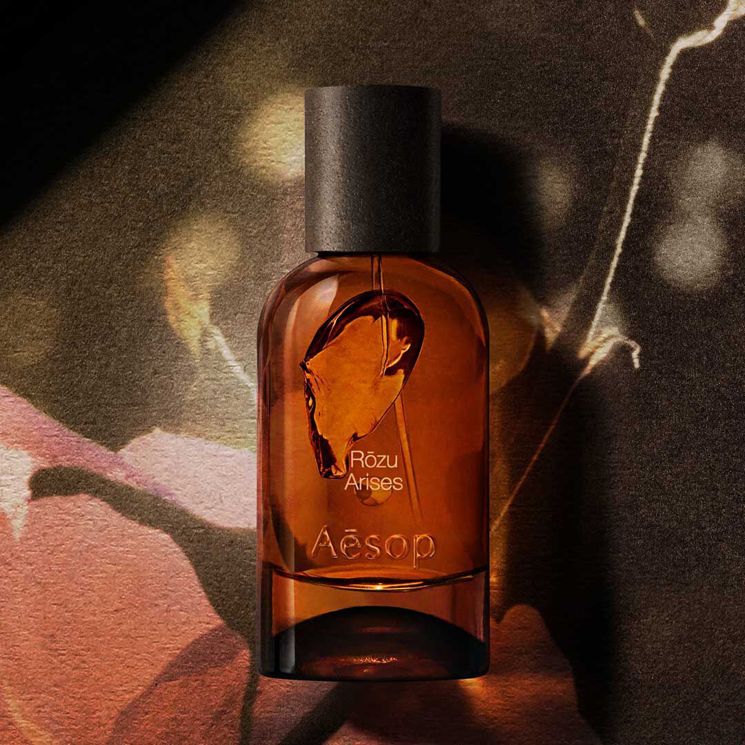 Frasco ambar de fragancia Aesop Rozu Arises sobre una imagen floral oscura, con luz calida y sombras.