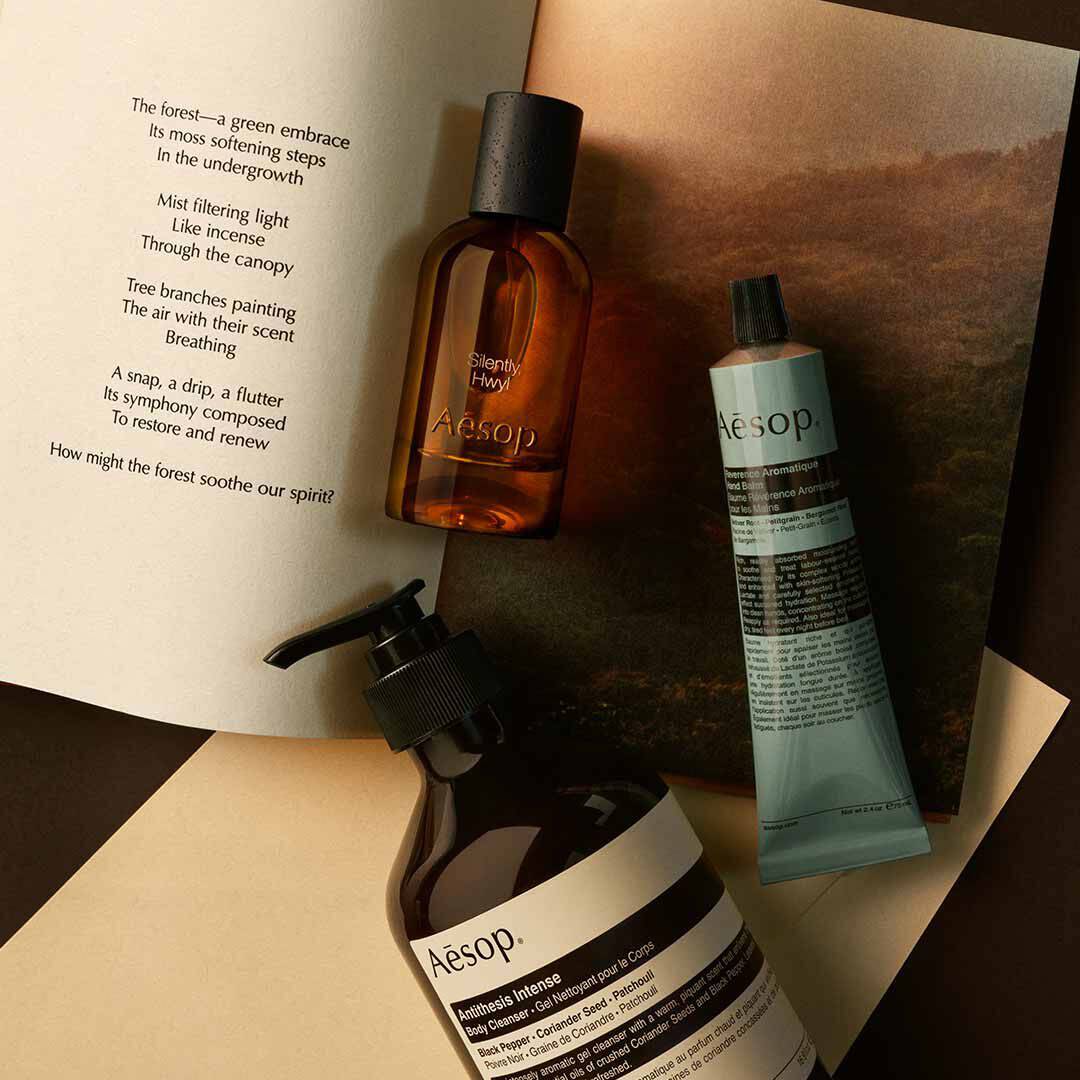 Frasco de Aesop Silently Hwyl con dispensador y tubo azul, dispuestos sobre papeles con una imagen de paisaje.