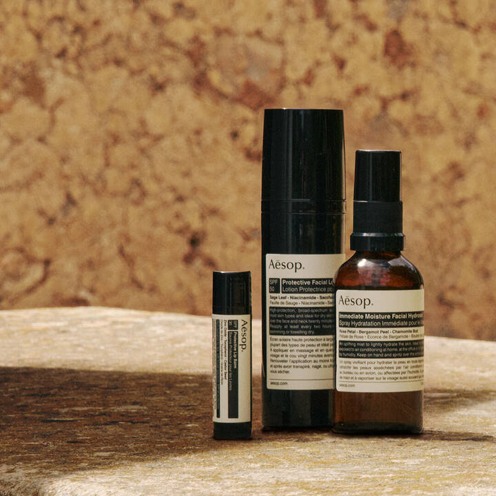 Una cuidada seleccion de formulaciones de proteccion solar de Aesop dispuestas sobre una superficie de piedra banada por el sol, contra una pared de tonos terrosos y calidos.