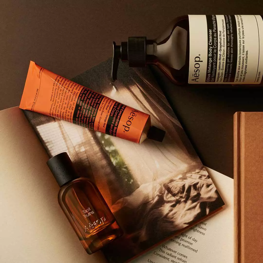 Frasco de Aesop Tacit Truths con dispensador y tubo naranja, dispuestos sobre papeles con una imagen monocromatica.