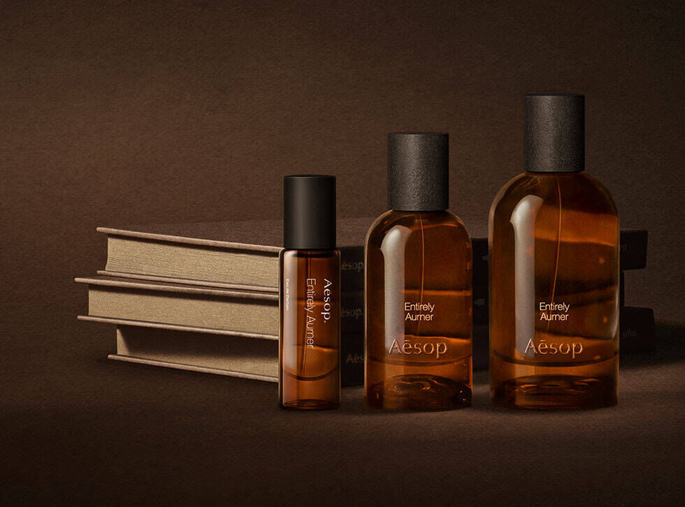 Tres frascos ambar de fragancias Aesop dispuestos junto a libros color crema apilados sobre una superficie marron oscura.