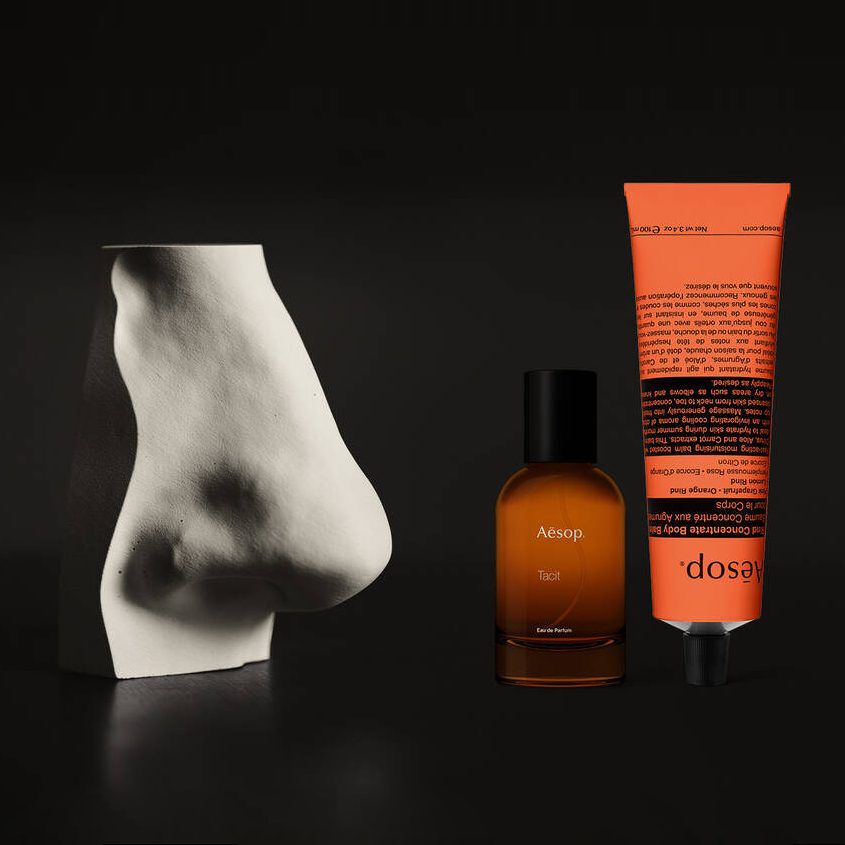 Frasco de perfume ambar y crema de manos coral junto a una escultura blanca