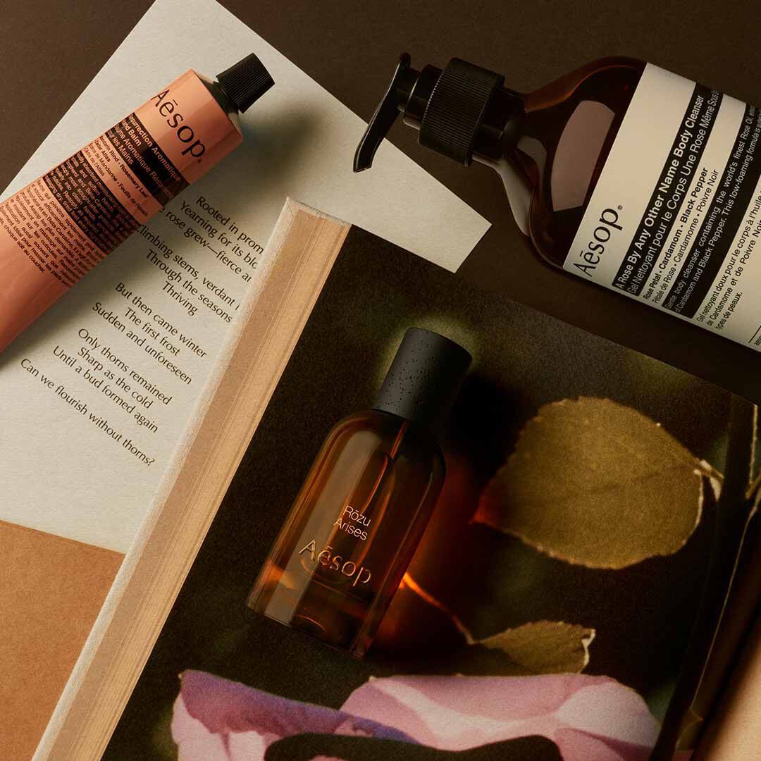 Frasco de Aesop Rozu Arises con dispensador y tubo color melocoton, dispuestos sobre papeles con petalos rosas y una hoja.