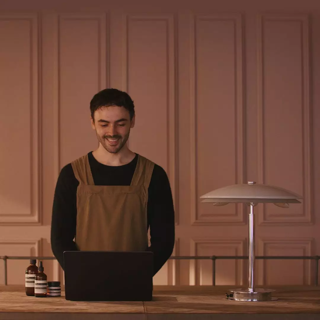 Una persona sonriente, con un delantal marron, mira un ordenador portatil detras de un mostrador de madera, junto a frascos de vidrio ambar y una lampara de mesa metalica.