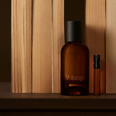 Botella ambar de Aesop y vial esbelto dispuestos entre tablas verticales de madera clara, con sombras marron oscuro y una composicion lineal depurada.