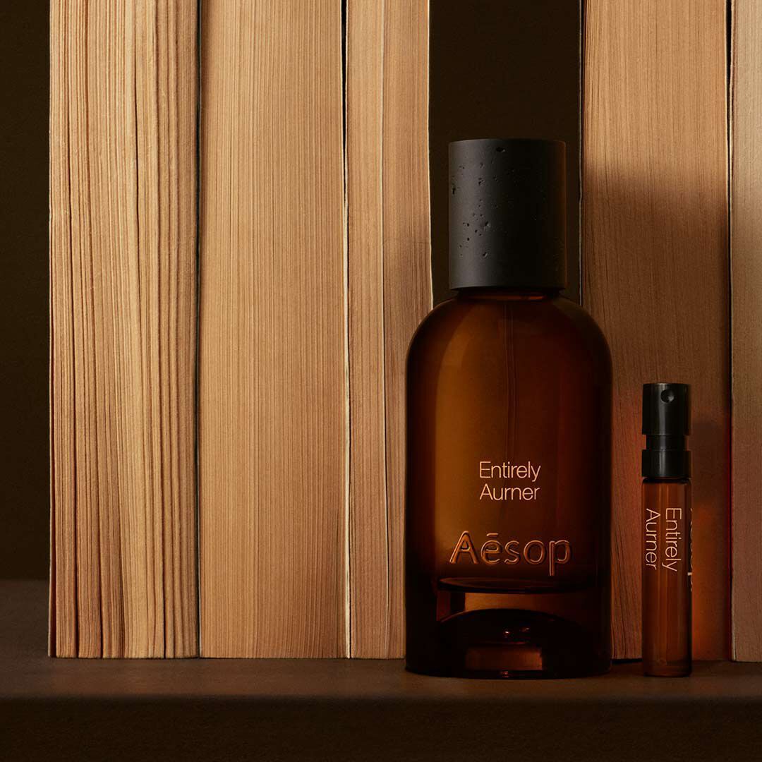 Un frasco de vidrio ambar del perfume Aesop Entirely Aurner y un vial de muestra a juego colocados en vertical junto a libros alineados sobre una superficie oscura.