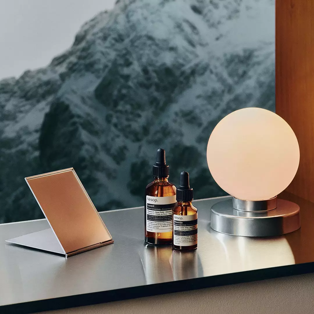 Two amber Aesop dropper bottles stand between a folding mirror and a glowing globe lamp on a reflective ledge, with snowy mountains beyond.