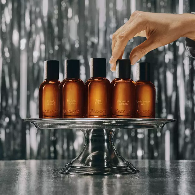 Una fila de frascos de perfume de vidrio ambar de Aesop exhibidos en un estante de madera frente a una biblioteca y una obra de arte verde.