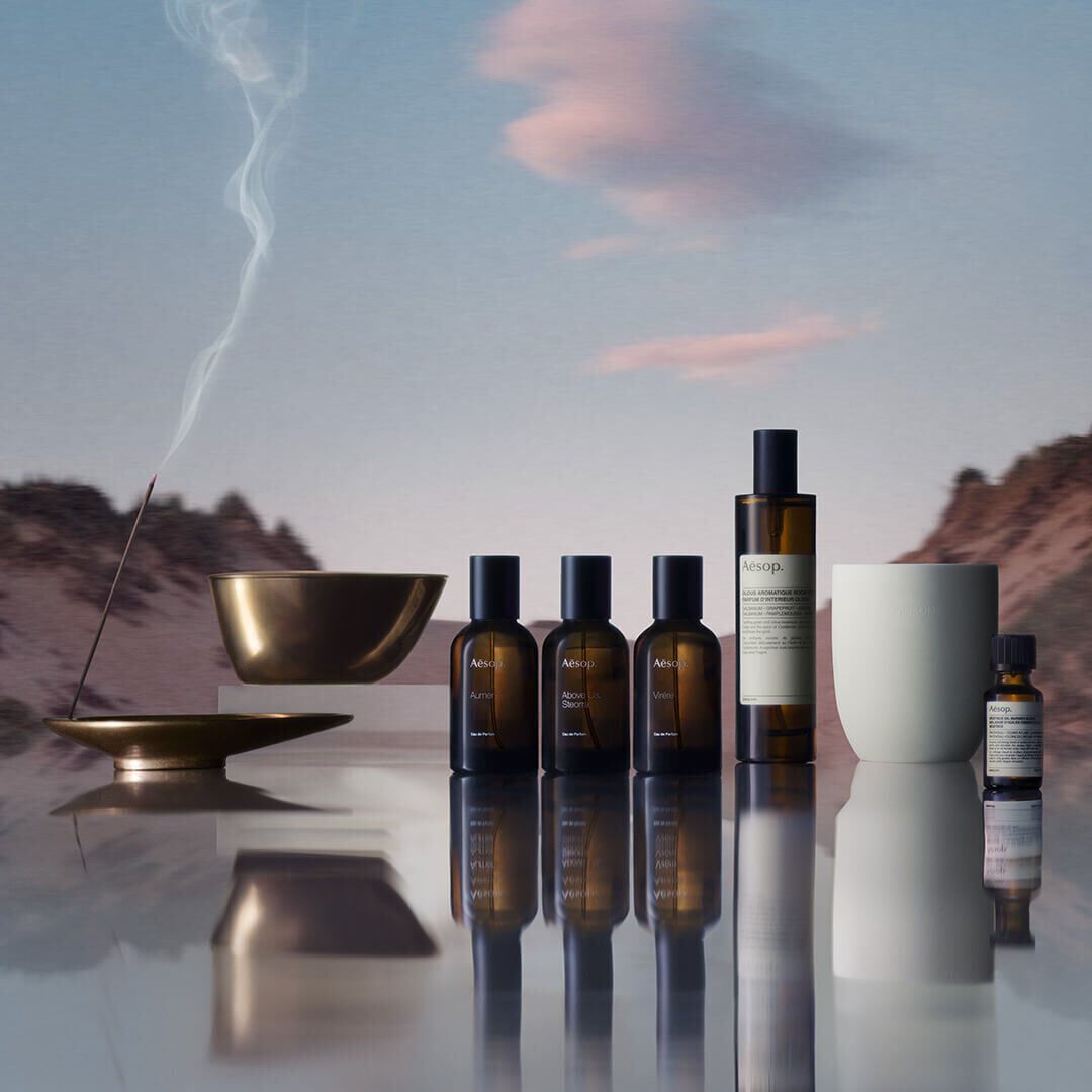 Una coleccion de productos Aesop, que incluye perfumes, ambientadores y aceites esenciales, con una varilla de incienso encendida en un soporte de bronce, todo ello con un paisaje montanoso difuminado reflejado en una superficie.