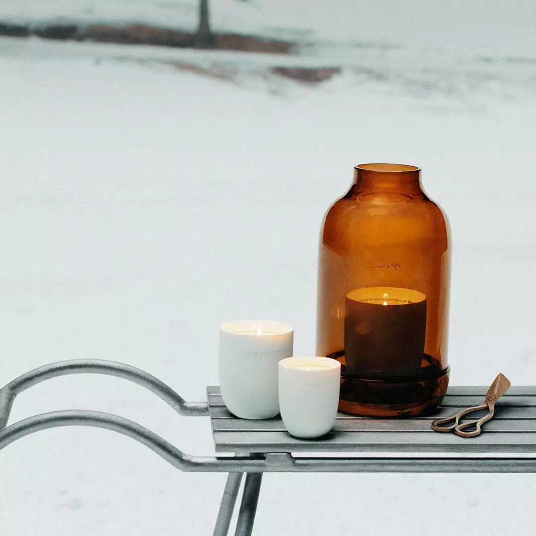 Two white candle vessels, an amber glass lantern containing a lit candle and wick trimmers sit on a metal sledge in snow.