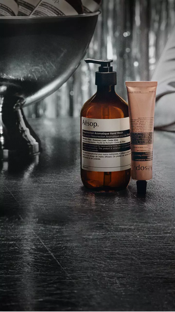 Una botella ambar Aesop con dosificador y un tubo rosa se alzan junto a un gran cuenco con pedestal sobre una mesa reflectante, con serpentinas plateadas al fondo.