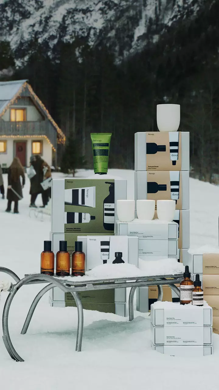 Cajas, botellas ambar, tubos de colores y recipientes blancos de Aesop dispuestos alrededor de un trineo de metal sobre la nieve, con personas frente a una casa iluminada al fondo.