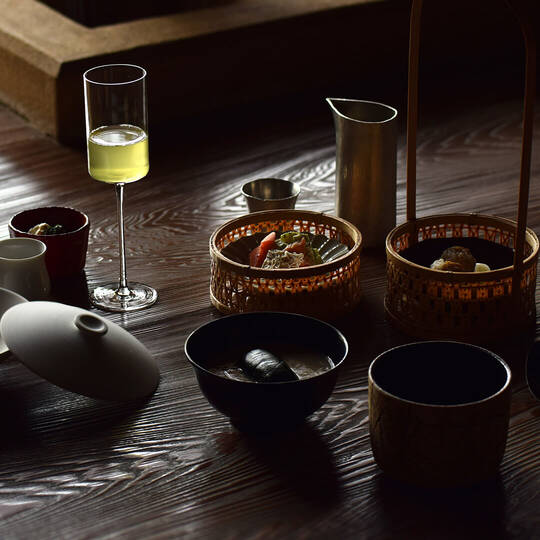 A dark, textured wooden table with various Japanese dishes and an elegant stemmed glass.