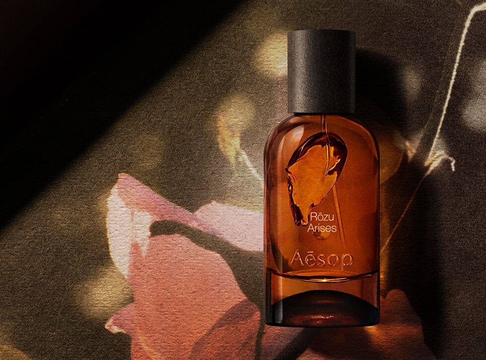 Frasco ambar de fragancia Aesop Rozu Arises sobre una superficie oscura, junto a una ilustracion botanica rosa y una raiz clara.