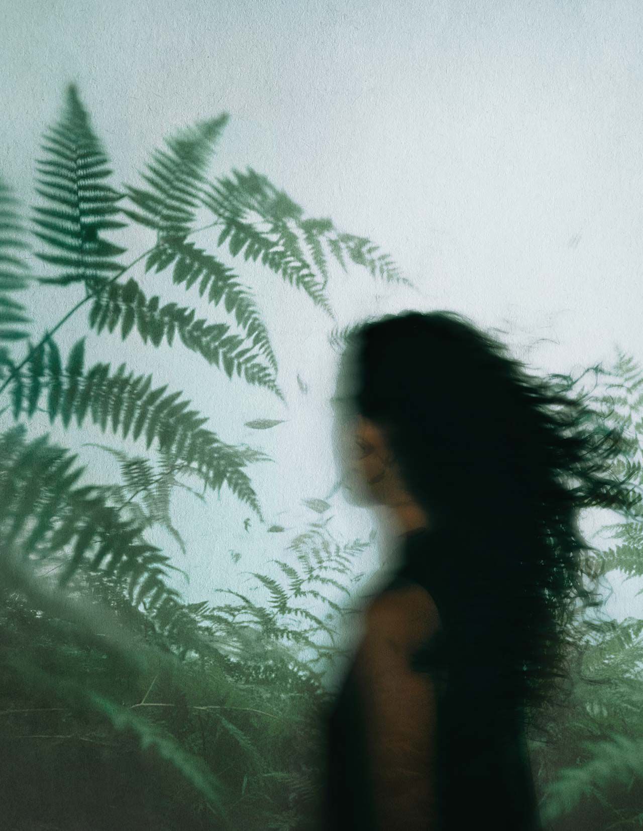 Perfil difuminado de una persona de cabello oscuro entre helechos verdes, sobre un fondo de vidrio empanado y luz natural fria y difusa.