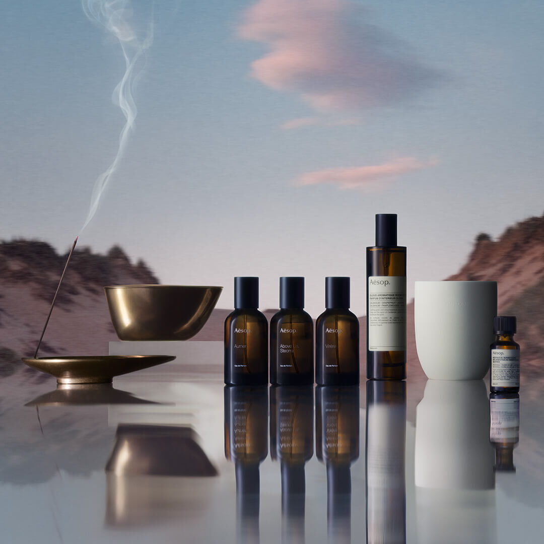 Una coleccion de productos Aesop, que incluye perfumes, ambientadores y aceites esenciales, con una varilla de incienso encendida en un soporte de bronce, todo ello con un paisaje montanoso difuminado reflejado en una superficie.