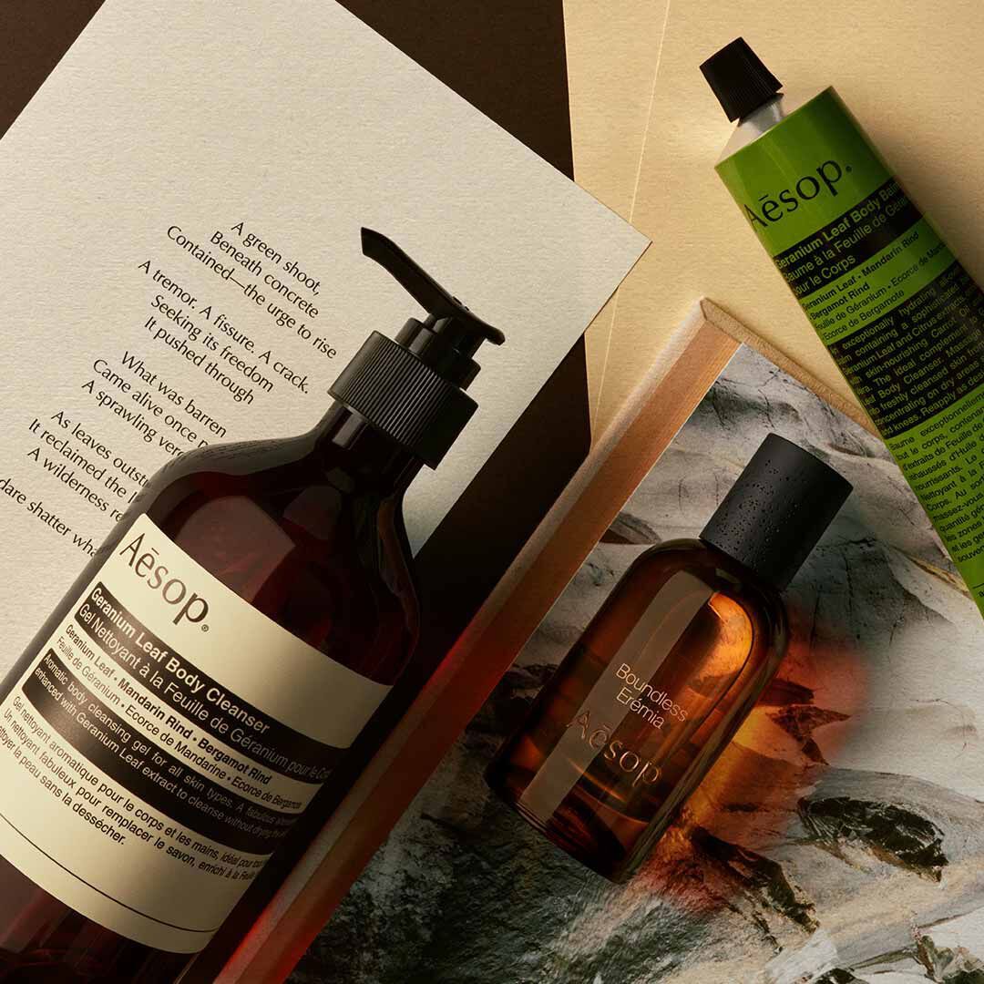 Frasco de Aesop Boundless Eremia con dispensador y tubo verde, dispuestos sobre papeles y una imagen de piedra.