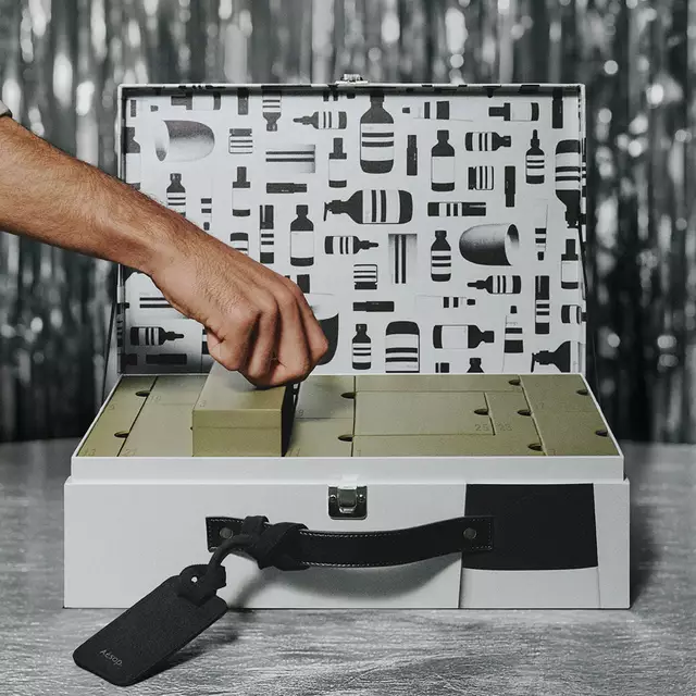 Una mano levanta una caja verde con compartimentos de un estuche plateado abierto con tapa estampada en blanco y negro, sobre un fondo de oropel plateado.