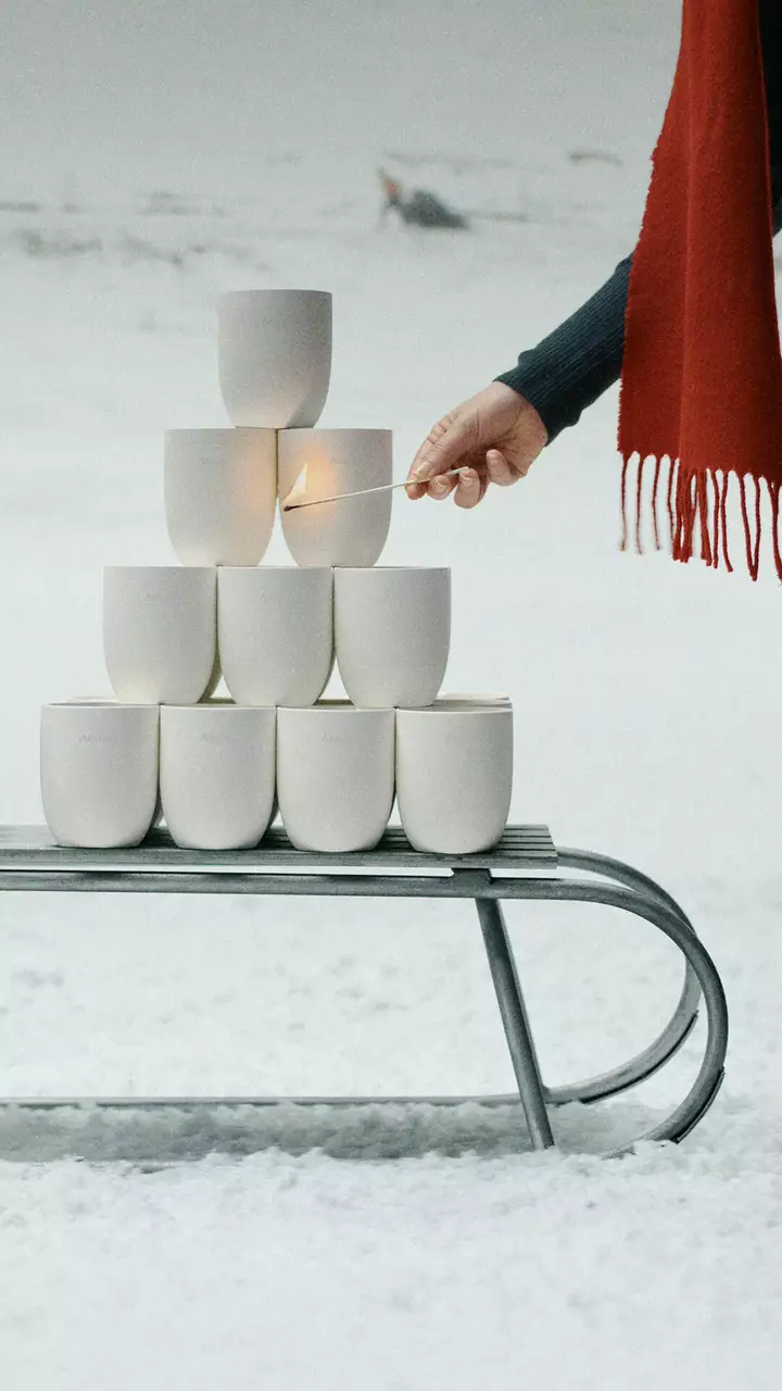 Una mano sostiene una cerilla encendida junto a recipientes blancos apilados sobre un trineo metalico en la nieve, con una bufanda roja con flecos a la derecha.